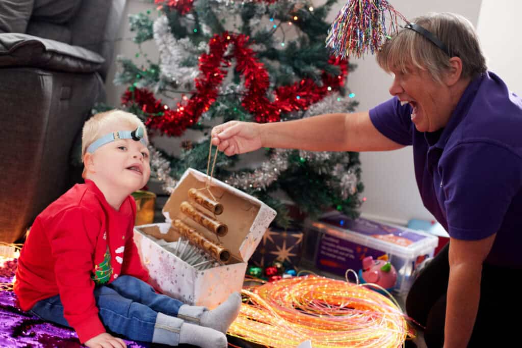 Arthur, a small boy wearing a Christmas jumper, playing in front of a Christmas tree with Sense support worker Nicky.
