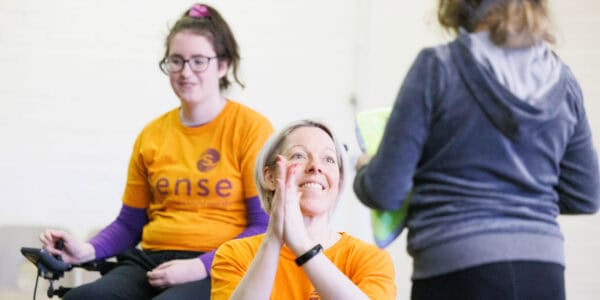 A woman wearing a Sense t-shirt claps her hands while leading a dance activity for people with disabilities.