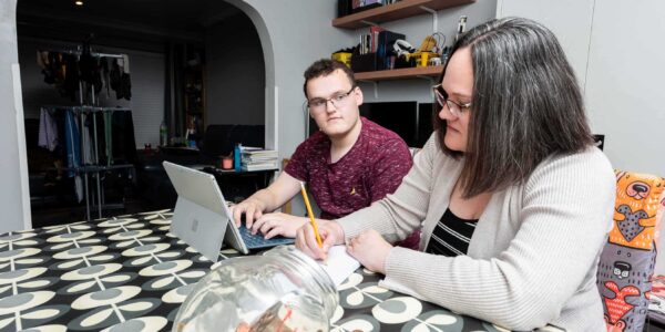 At a table, a woman with short dark hair and glasses is writing on a notepad. Next to her, a young man with short dark hair and glasses is looking concerned and typing into a computer.