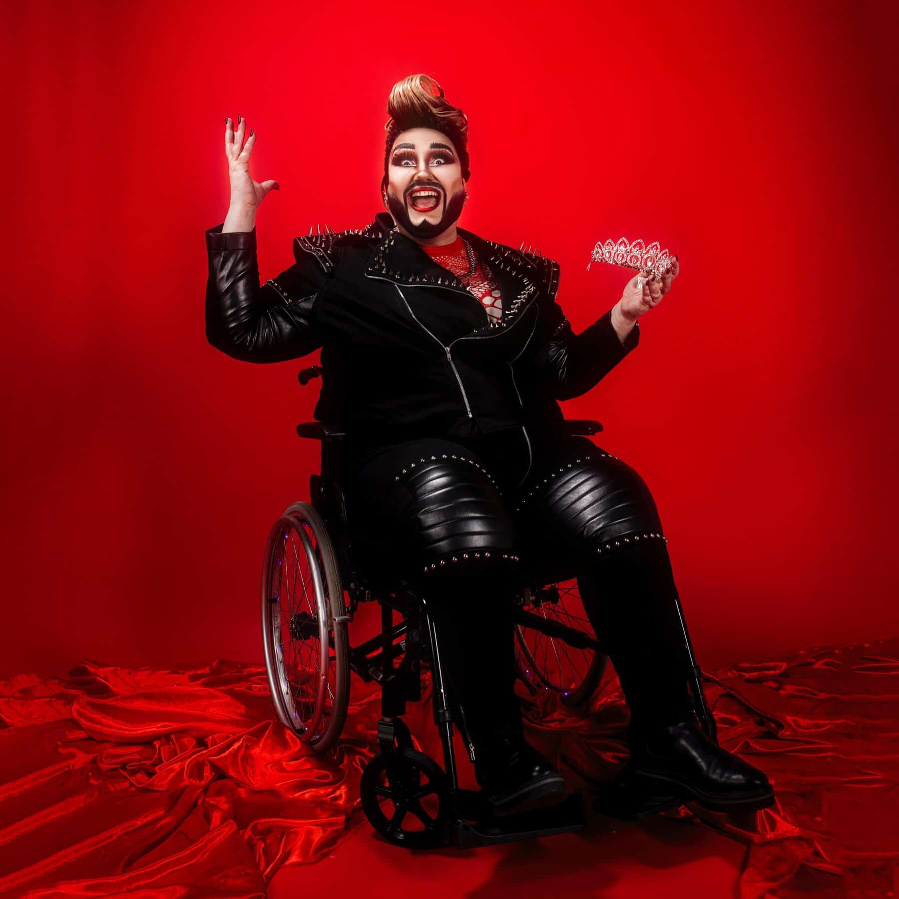 A drag artist sits in their wheelchair in a red satin background, holding a tiara in their left hand.