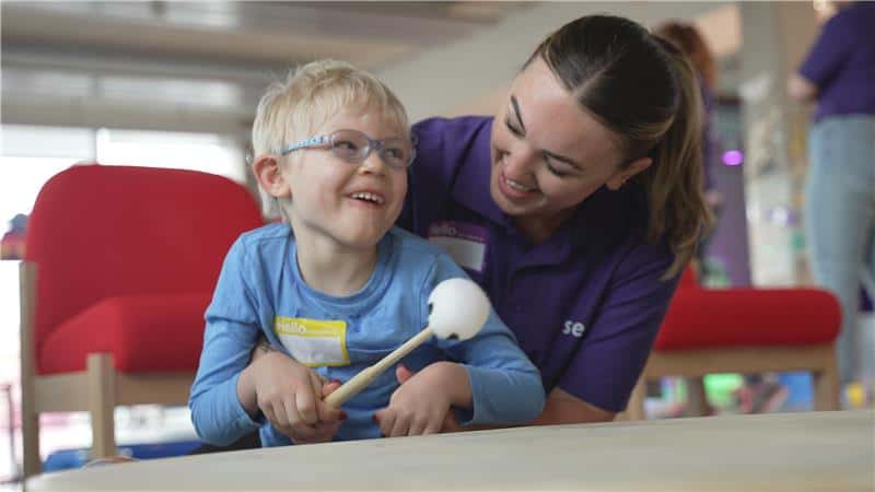 A boy using a drumstick on a drum assisted by a teacher.