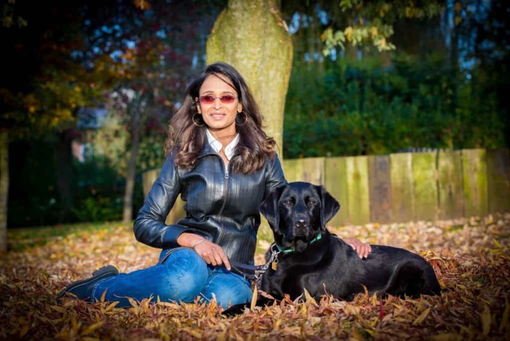 A woman with long hear, wears sunglasses and leather jacket, sitting with her dog.