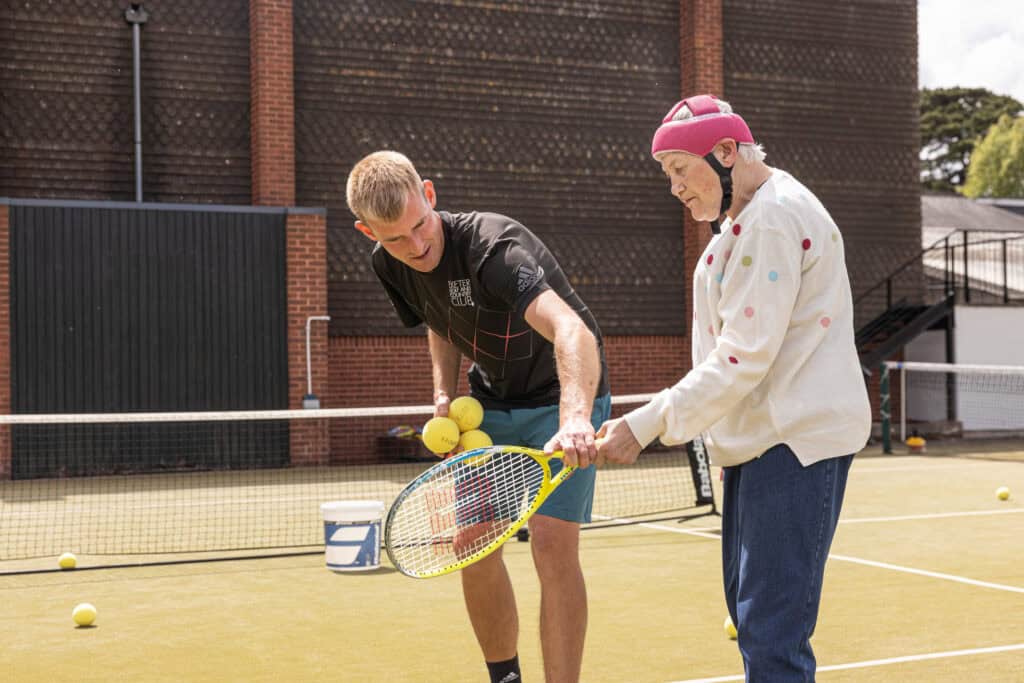 A woman wearing a white jumper holds a tennis racket whilst a young man with blonde hair puts his arm towards the racket. They are on a green tennis court with a net in the background.