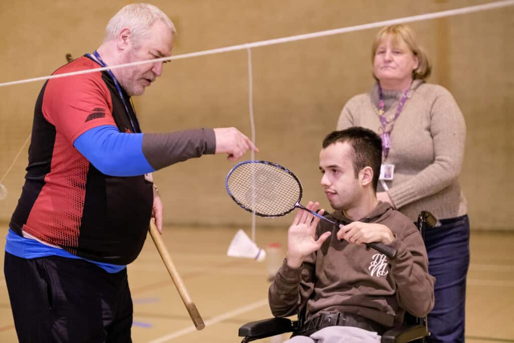 A boy with brown hair sits in a wheelchair holding a badminton racket. A woman stands behind him and a man with white hair holds a shuttlecock out for him to hit.