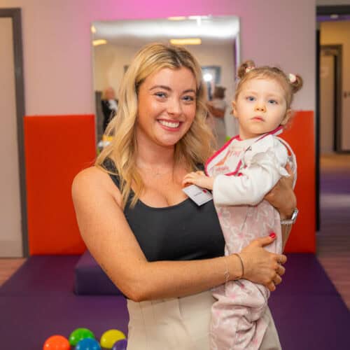 A young woman is smiling to camera, she's holding a small girl in her arms