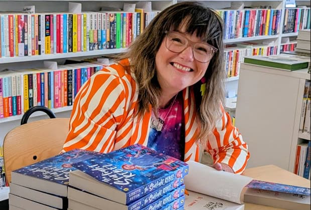Lisette Auton leaning over her new book, The Starlight Rebel. She is smiling widely and wearing an orange and white striped shirt.