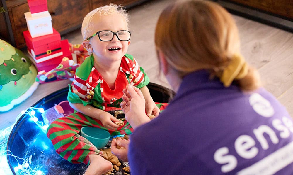Harvey is wearing glasses and sat in his elf suit, inside a messy play area. He is smiling widely back at the Sense employee.