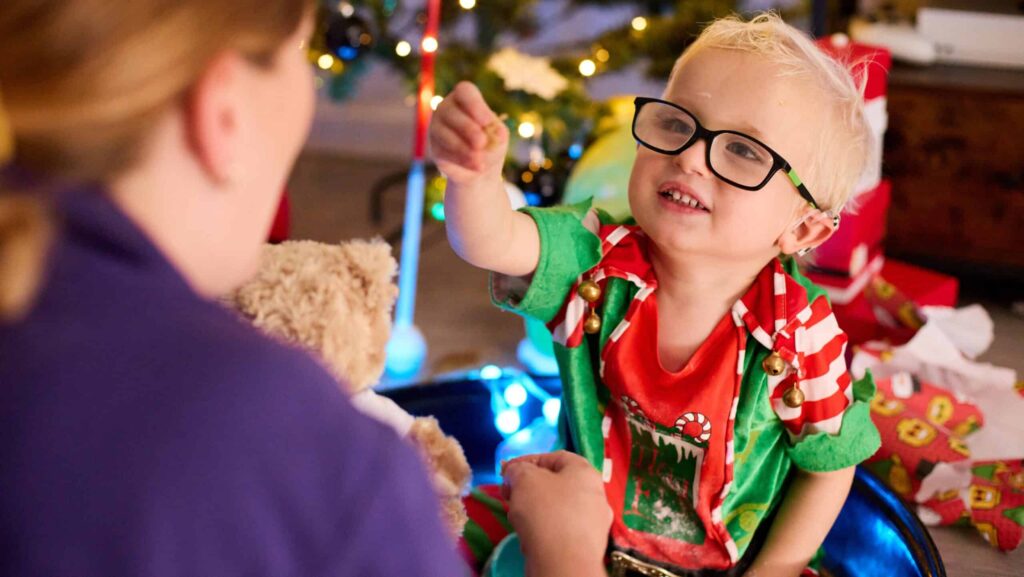 Harvey is sat down in his elf suit, leaning forward showing off an item to Laura in front of him.