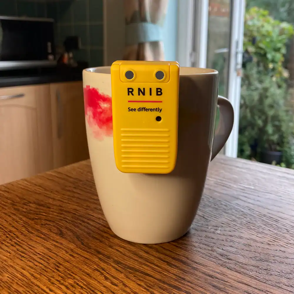 Yellow RNIB liquid level indicator clipped to the side of a white mug on a wooden table, used to help blind and partially sighted people detect when a cup is nearly full.