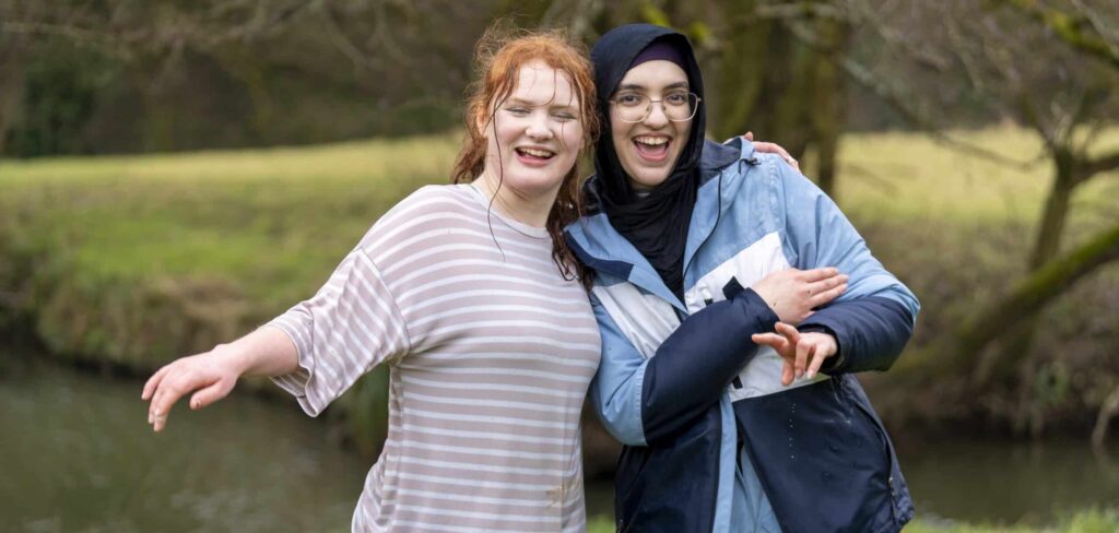 Rumaysa and clementine are stood side by side, smiling at the camera. They are both sodden after jumping in the river behind them.