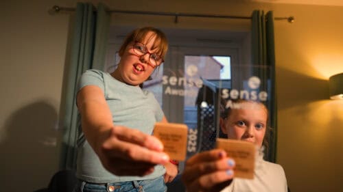 Two girls pose in their living room. They are holding up wooden and glass trophies which read Sense Awards