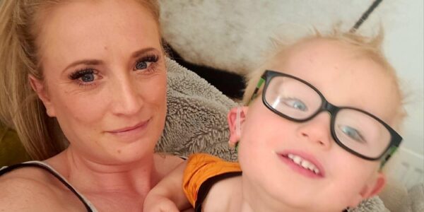 A young boy wearing glasses smiles at the camera next to his mum, who has blonde hair and is wearing a grey t-shirt