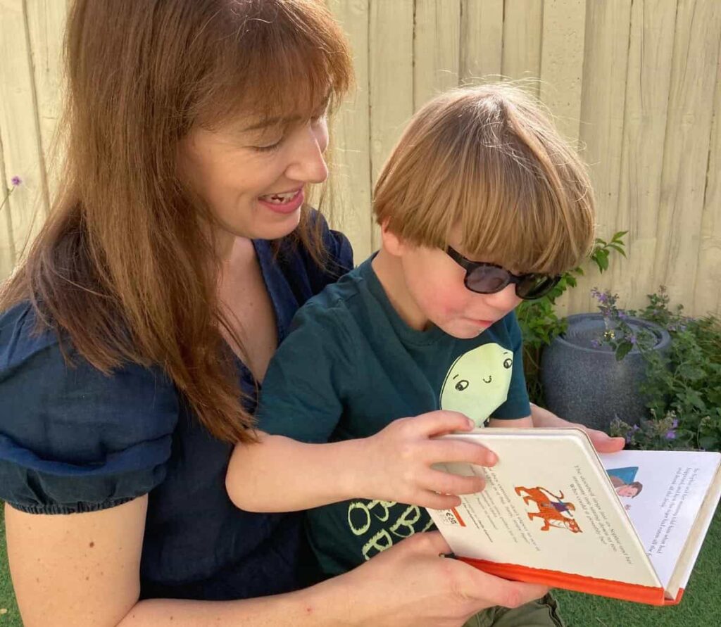 Sonny is sat on Julie's lap, holding and reading a book. He is wearing dark glasses and looking down at the book concentrating. Julie is smiling wearing a dark blue dress.