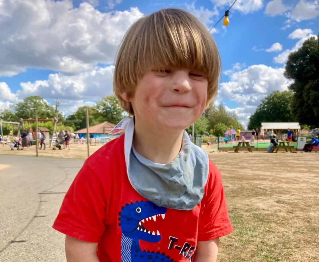 Sonny is stood in a park, wearing a red t-shirt with a blue T-Rex on it. He is smiling widely at the camera.
