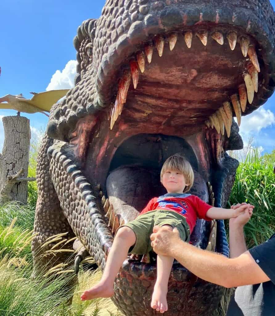 Sonny is sat in the mouth of a huge model T-Rex, who has its jaw wide open. He is posing for a picture and smiling back at the camera in his red T-Rex t-shirt.