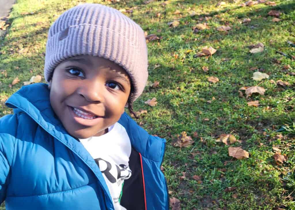 Azuriah is in a green field, covered in brown leaves. He has a blue jacket coat and a pink beanie on. He is smiling widely and his eyes are big and brown.