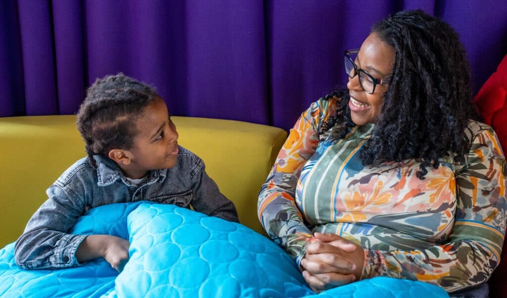Natalie and Azuriah are sat on a yellow sofa, with a big blue bean blanket across both of their laps. They are looking at each other and smiling widely.