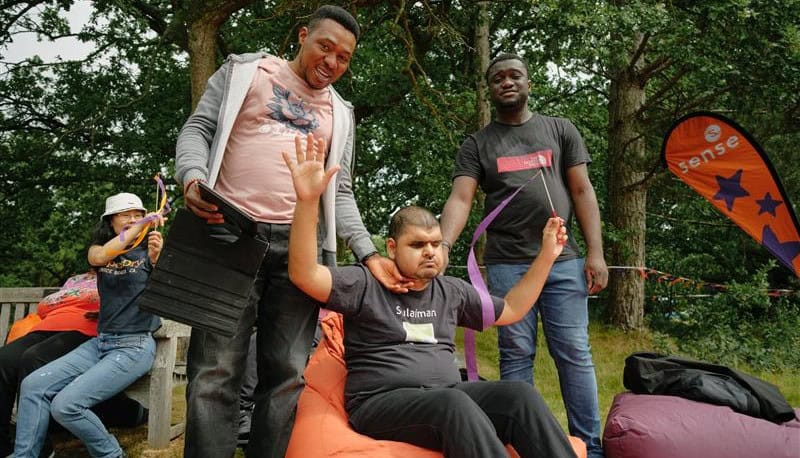 Three men are posing for a picture in a park. Two support workers are stood either side of a man who is sat on a beanbag with his hands in the air, holding a purple ribbon in his left hand.