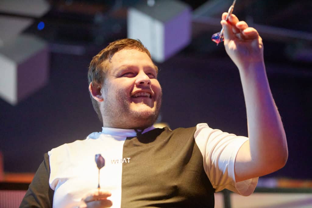 Ryan has a dart in his left hand. His arm is bent at a ninety degree angle, getting ready to throw the dart. He is smiling widely.