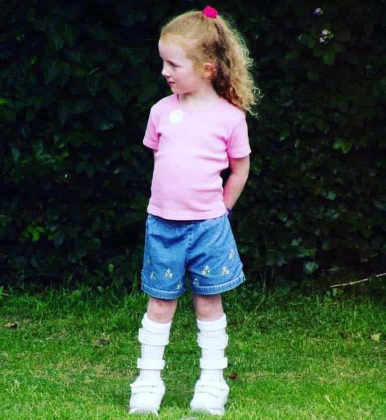 Siobhan at age six, stood on grass wearing a pink top and blue jean shorts. She has a custom brace around her legs to help her walk.