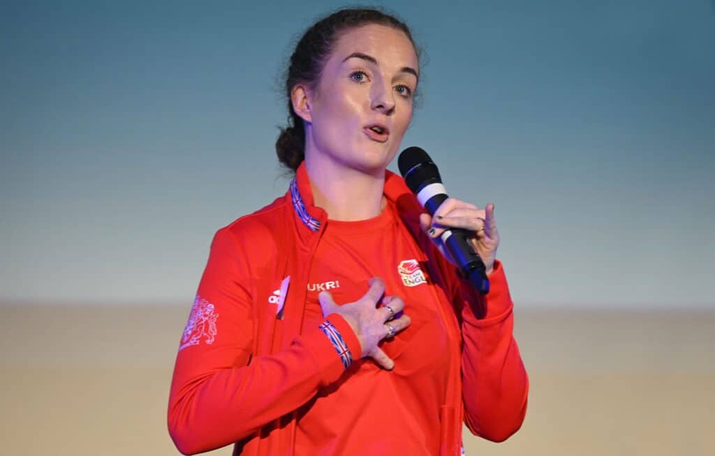Siobhan is stood in a red Team England jacket, holding a microphone to her mouth. She is speaking on a stage.