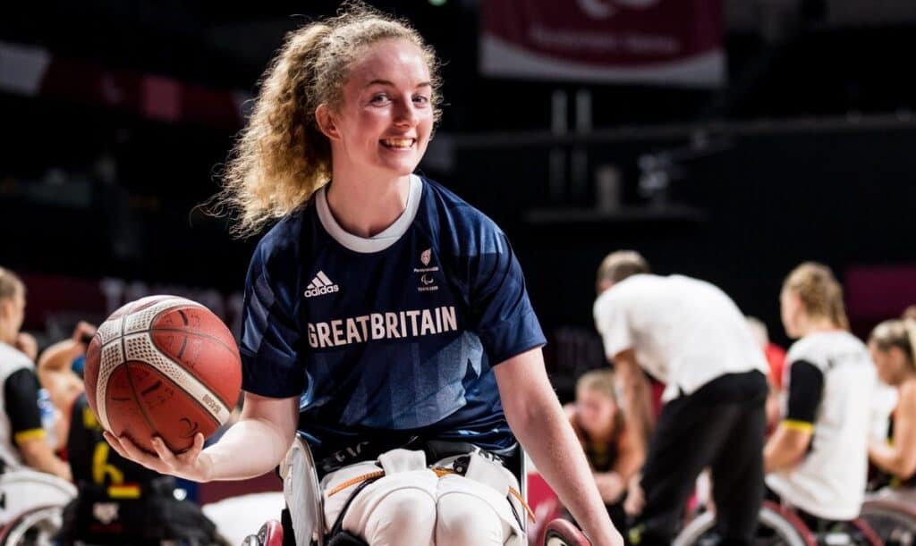 Siobhan is sat in her wheelchair, with a basketball in her right hand. She is smiling widely at the camera, and wearing a sports top that reads 'Great Britain'