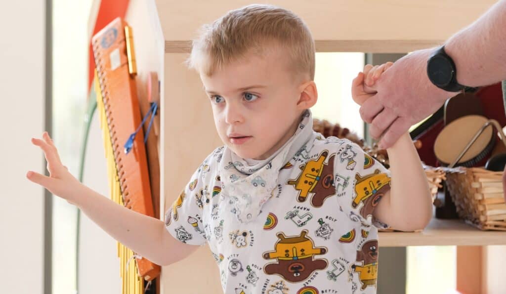 Thomas is stood, holding his dads hand for balance. He is facing the camera with a concentrated look on his face and is wearing a white t-shirt and neck tie with bears on it.