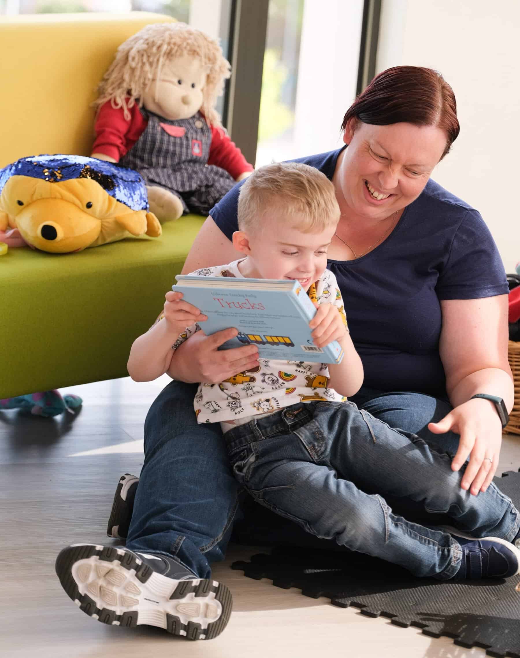 Thomas is sat on Caroline's lap, and she is sat on the floor. They are both laughing and reading a book called 'Trucks'.