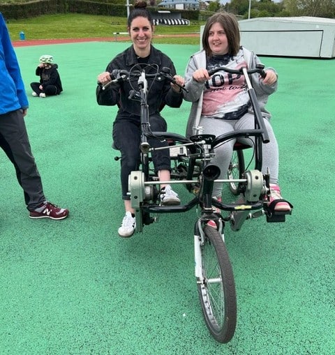 Chloe is sat on a side by side tandem bicycle. She and her friend are smiling widely.