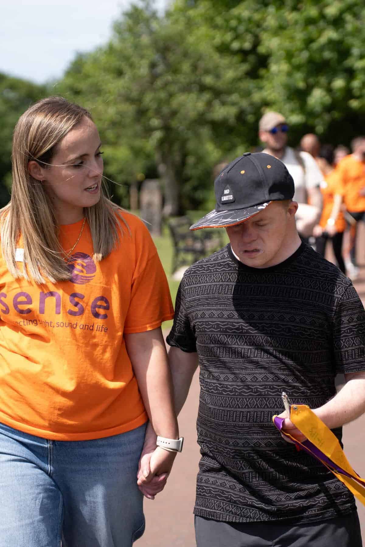 Corey is walking alongside Laura in an outdoor event. Corey is holding Laura's hand with his right hand and holding an orange and purple ribbon with his left hand.