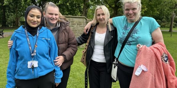 Four women are stood side by side in a park, all smiling widely.