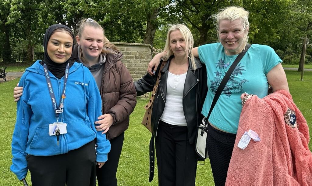 Four women are stood side by side in a park, all smiling widely.