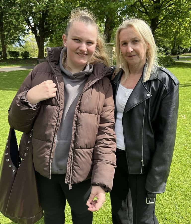 Kelsey and Paula are stood side by side in a park. It is sunny and they are smiling.