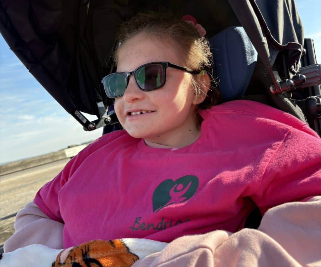 Megan is sat in her wheelchair in a pink jumper. She is smiling widely.