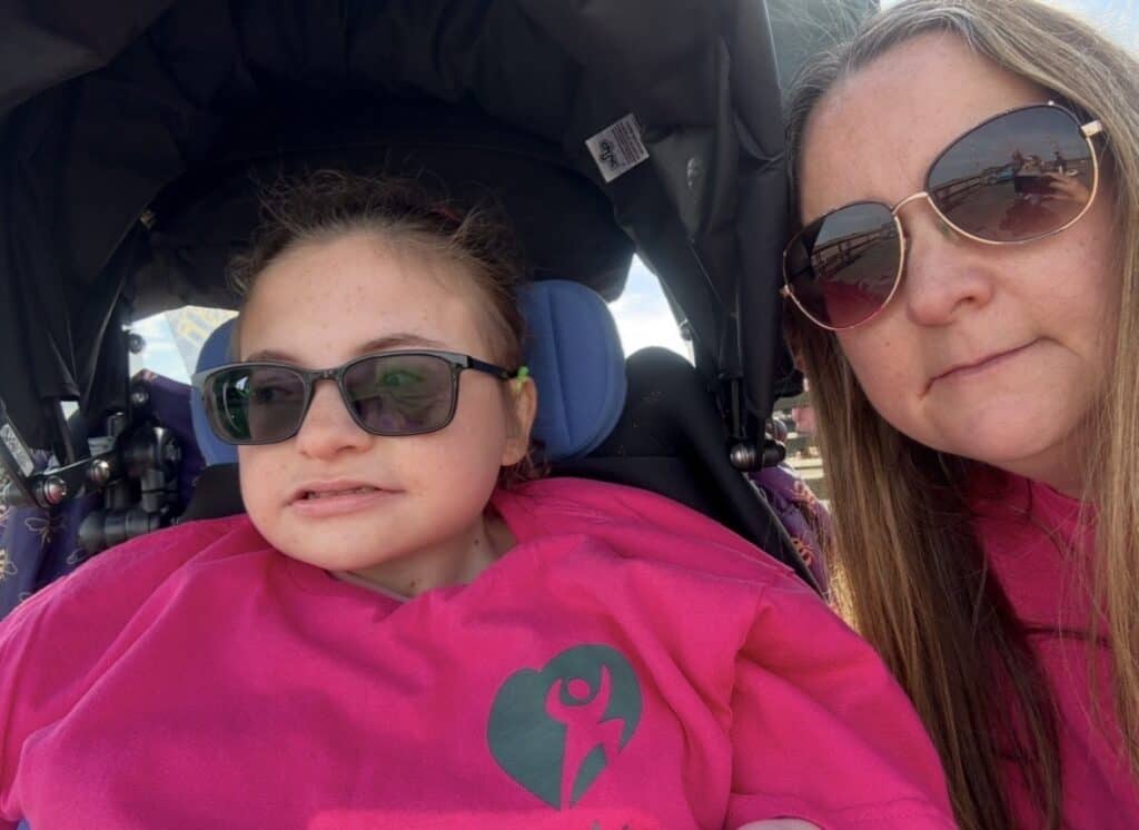 Megan is sat in her wheelchair in a pink jumper, her mother is posing next to her.