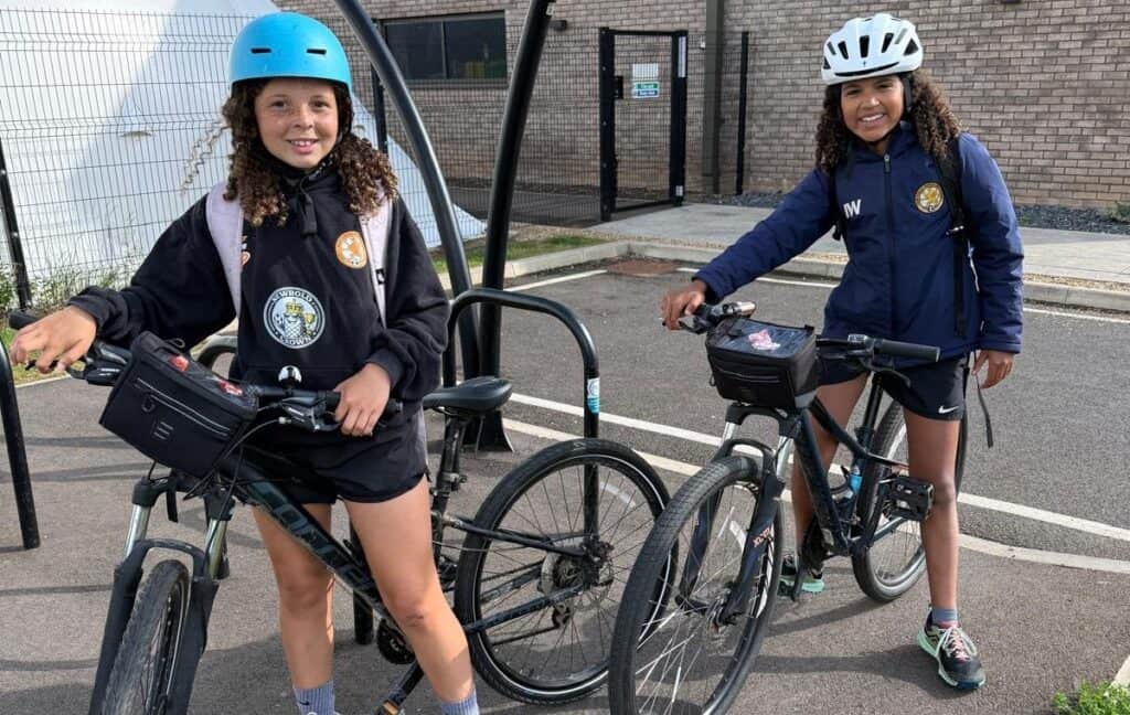 Esmae and Iris are in a car park, sat on their bikes. Esmae wears a blue helmet and Iris is in a white helmet.