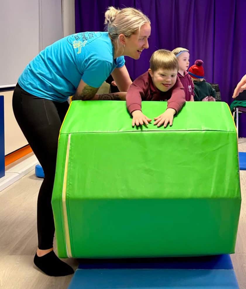 Freddie is about to tumble over a large green foam obstacle. Ella is supporting his legs from behind as they both smile widely.