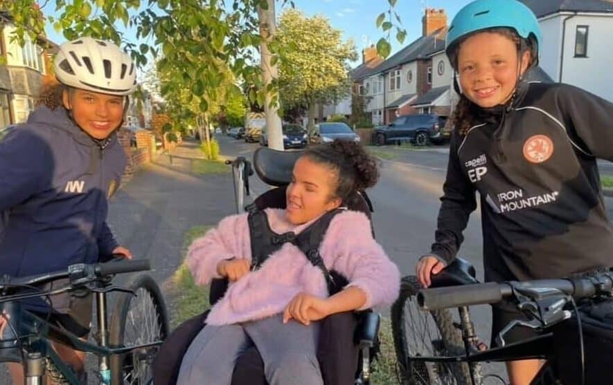 Iris and Esmae are stood either side of Mia. Iris and Esmae have their bikes in hand, and have helmets on for their training ride.