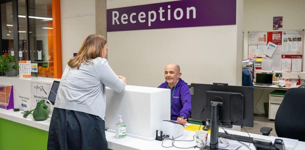 Joe is sat behind the reception desk, smiling widely, as someone leans over the desk to talk to him.