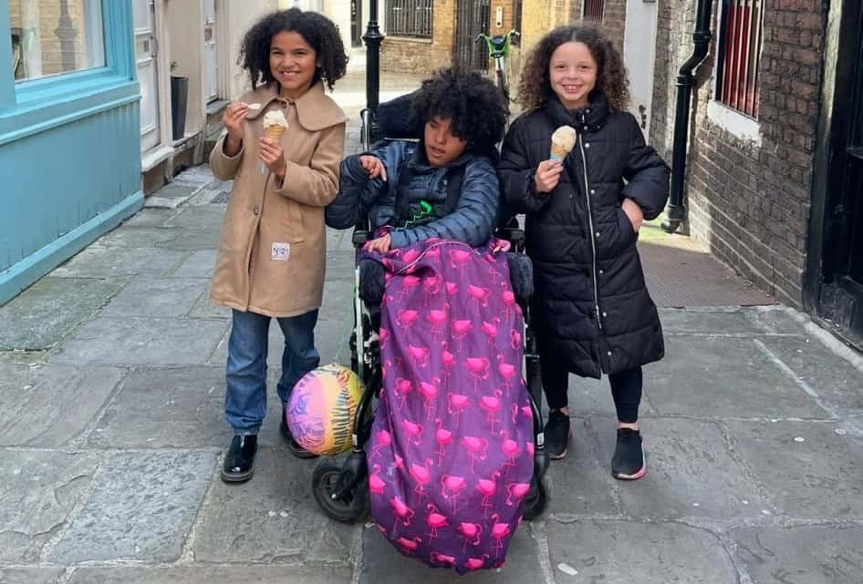 Iris and Esmae are stood either side of Mia. Mia is in her wheelchair. They are smiling widely and eating ice cream in a town setting.