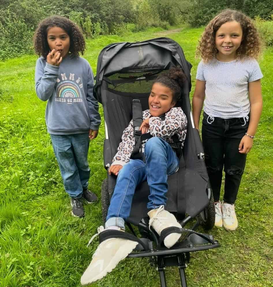 Iris and Esmae are stood either side of Mia. Mia is in her wheelchair. They are smiling widely in a luscious green park.