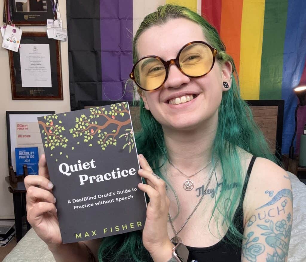 Max is sat on their bed, smiling widely and holding up their book titled 'Quiet Pratice: A DeafBlind Druid's Guide to Practice without Speech'.