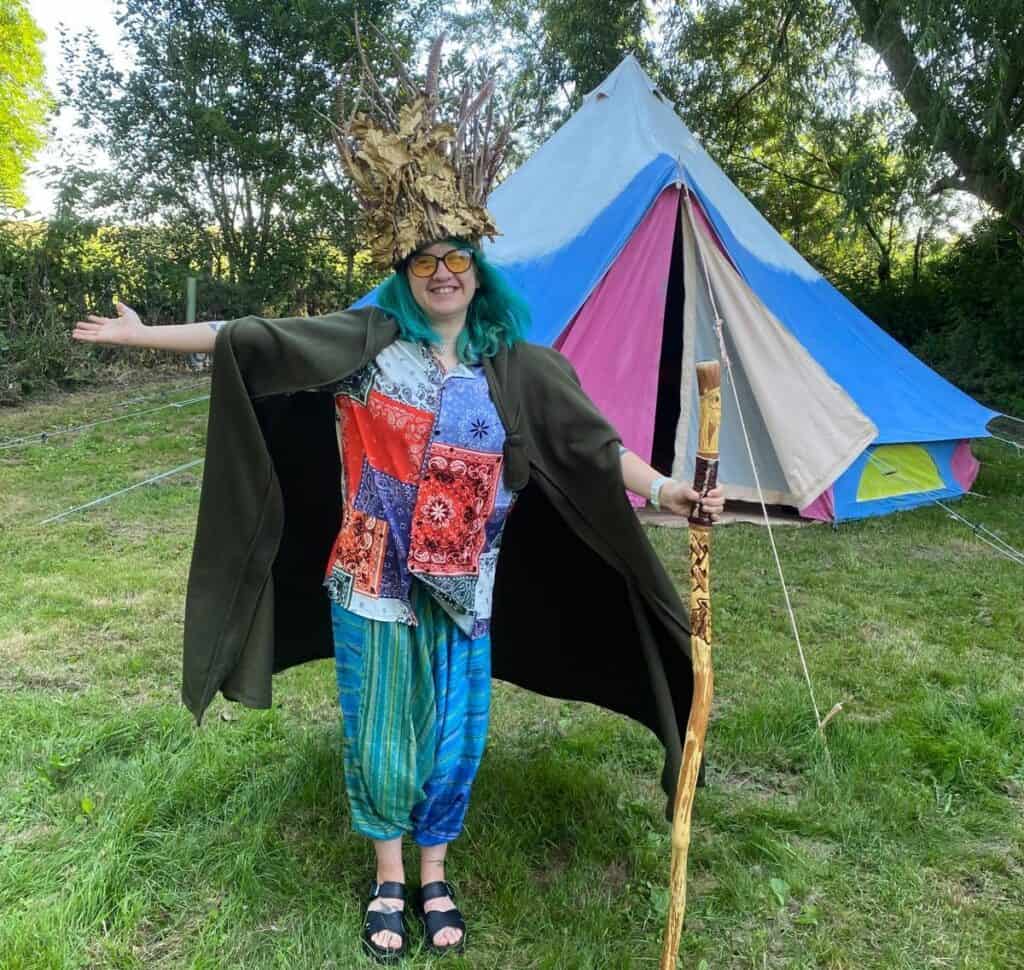 Max is stood in a colourful outfit, wearing a big green cape and a large hat made out of leaves and feathers. They are holding a wooden staff and smiling widely in front of their multi-coloured tent.