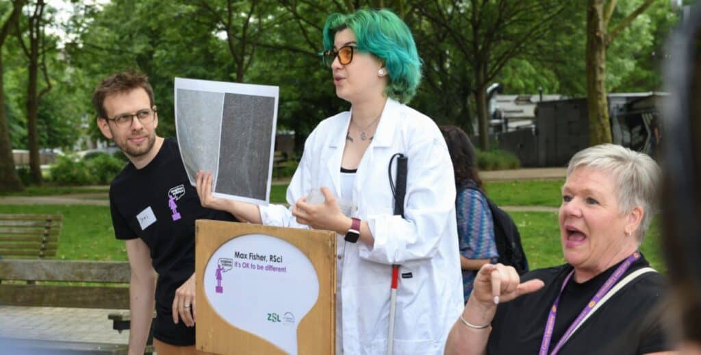 Max is stood behind a sign that reads 'Max Fisher - It's OK to be different', in a park, speaking to a crowd.