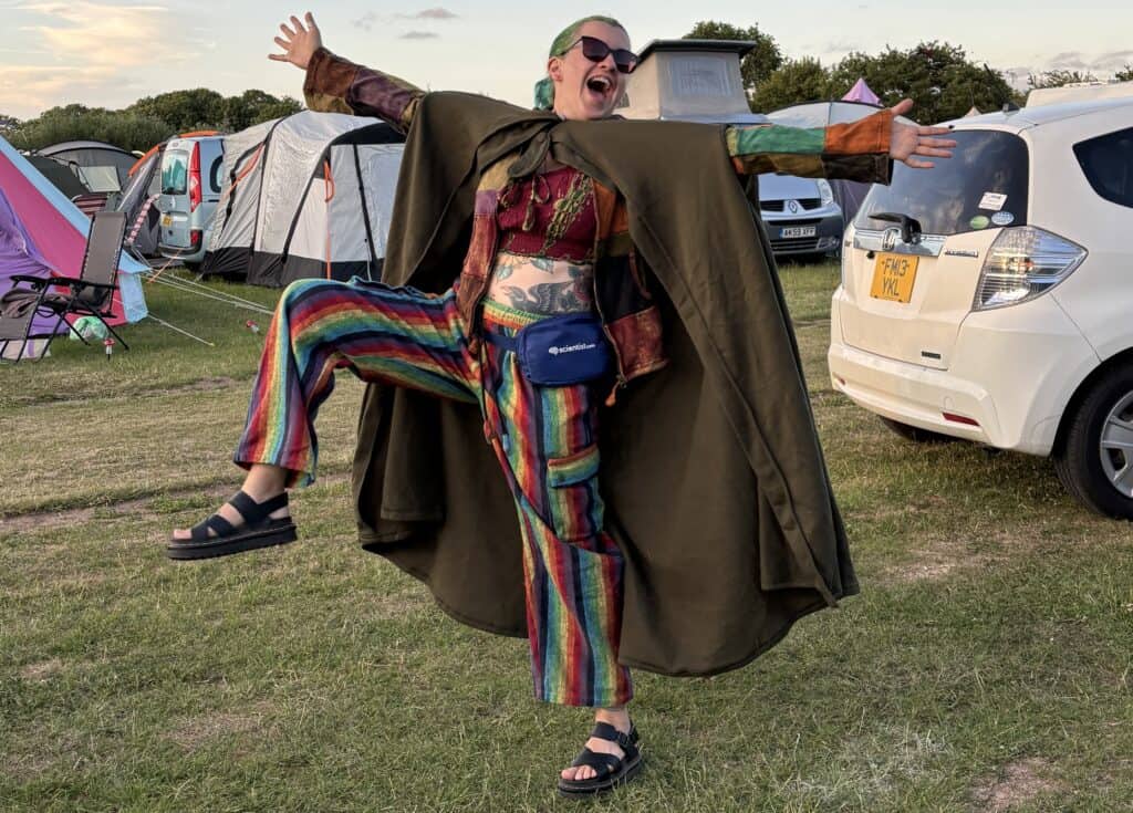 Max is balancing on their left foot, spreading their arms and right leg widely in a celebratory pose. They are wearing rainbow coloured trousers and a big green cape.