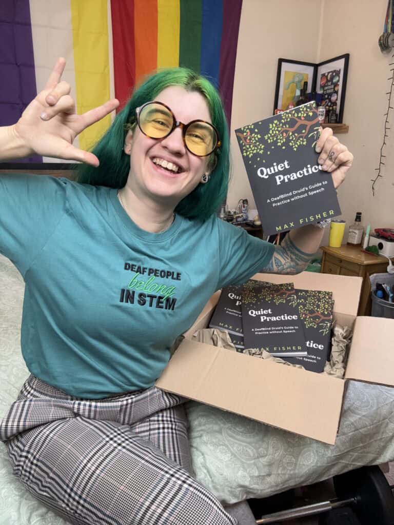 Max is sat on their bed, smiling widely and holding up their book titled 'Quiet Pratice: A DeafBlind Druid's Guide to Practice without Speech'.
