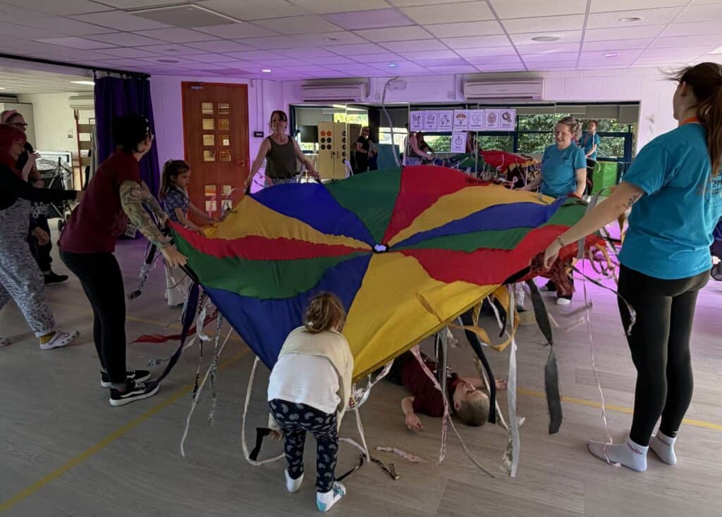 A group of people are playing with a multi-coloured parachute. They are stood in a circle and holding it over a young man, who is lying underneath it.