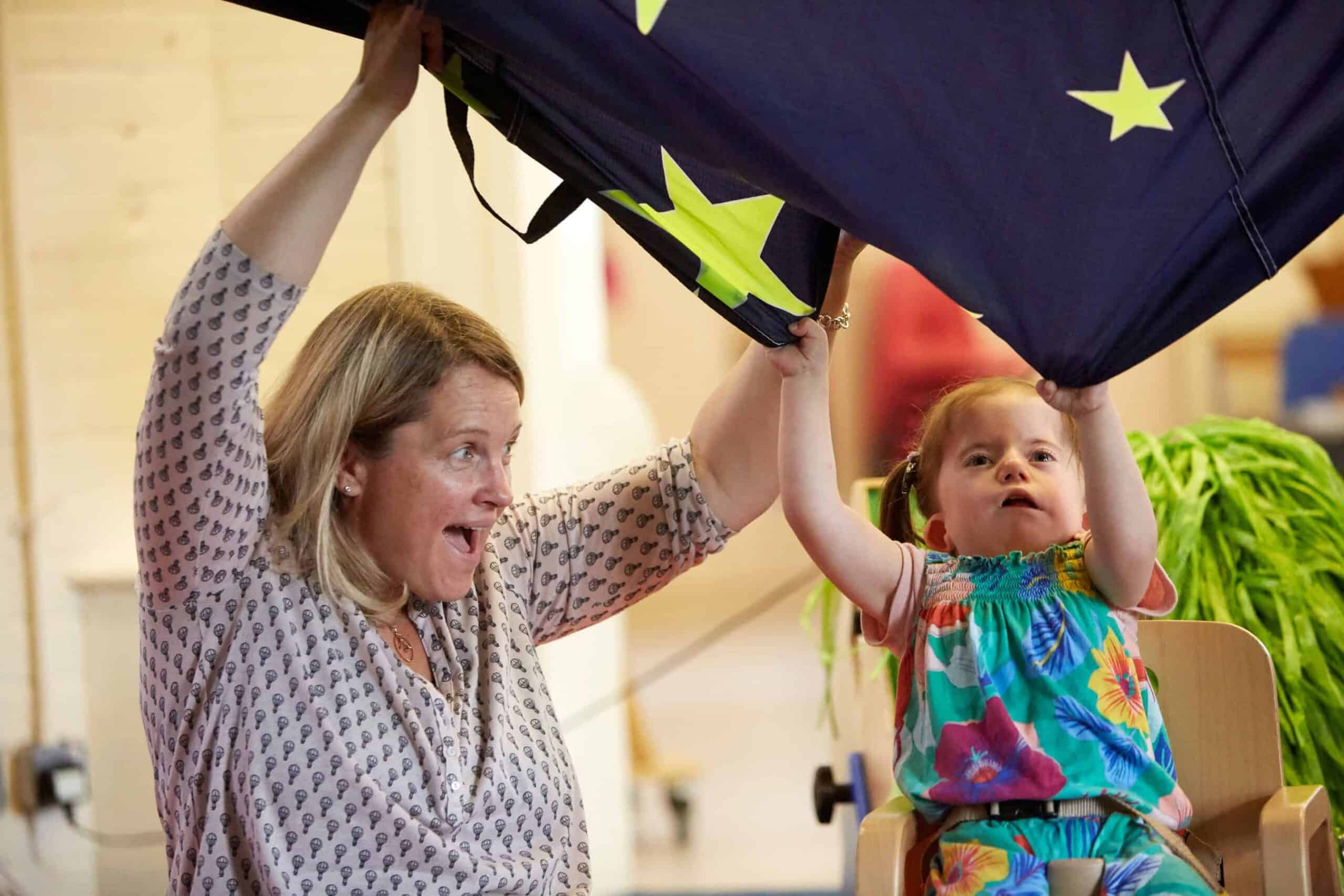 A young girl and a woman with blonde hair sit next to each other. Their arms are raised in the air as they wave a navy blue and star print parachute in the air.