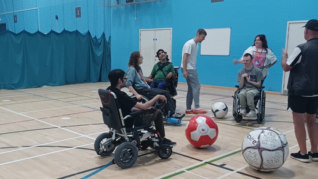 A group of adults sat in wheelchairs, in a sports hall, playing sensory football. There are three big inflatable footballs on the floor between them.