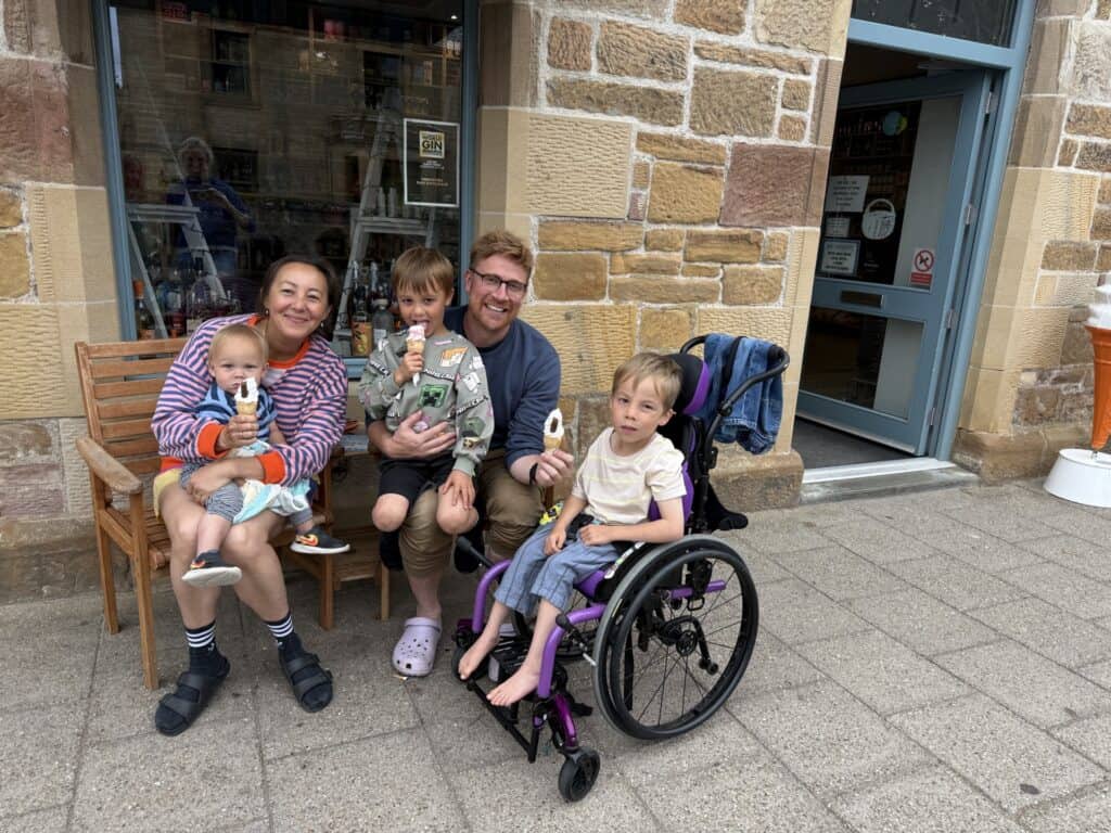 A family sitting on a bench, smiling and eating ice creams. There is a mum and dad, a baby boy, and two twin boys, one of whom is sitting in a wheelchair.